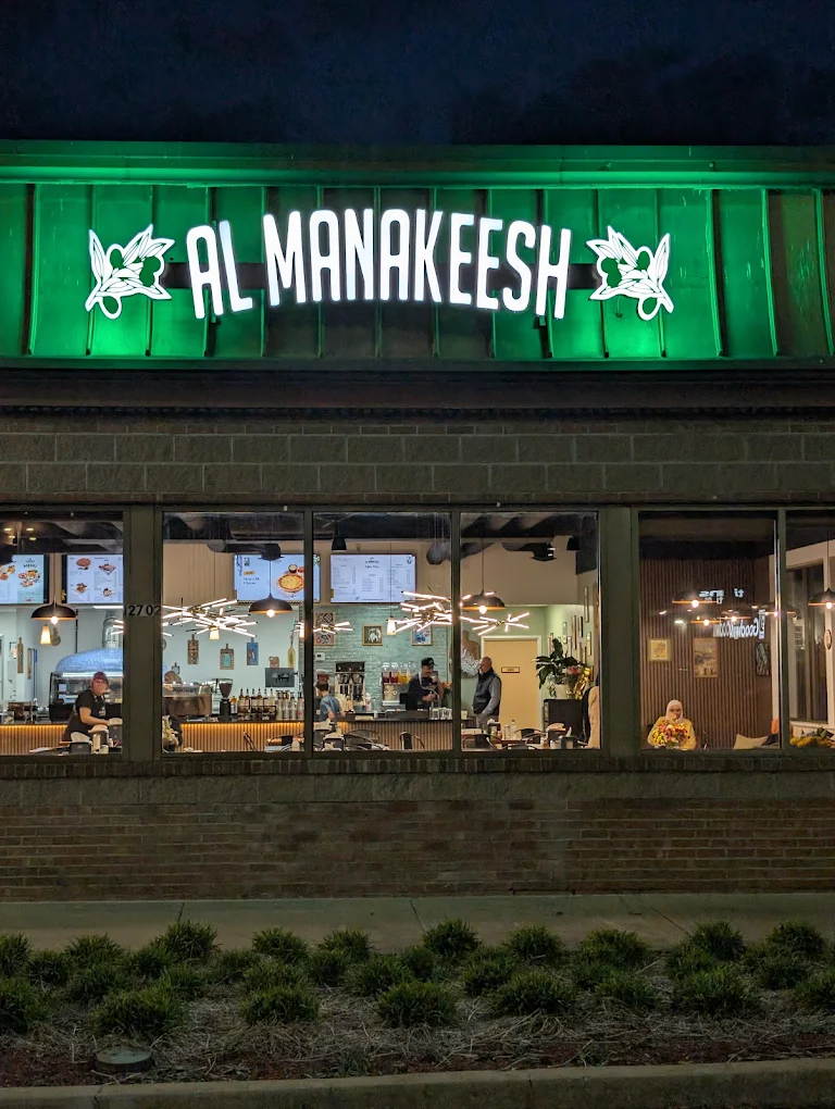 Al Manakeesh at night: a green backlit storefront sign above the dining room, with guests and the service line visible through full-height glass.